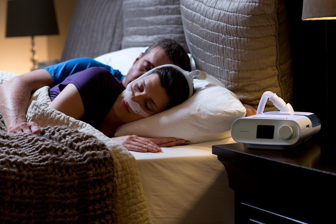 Philips Respironics Philips DreamStation Auto + Mask Package + Modem + Bonus Accessories, a woman sleeping with a CPAP mask on her face in a bedroom setting.